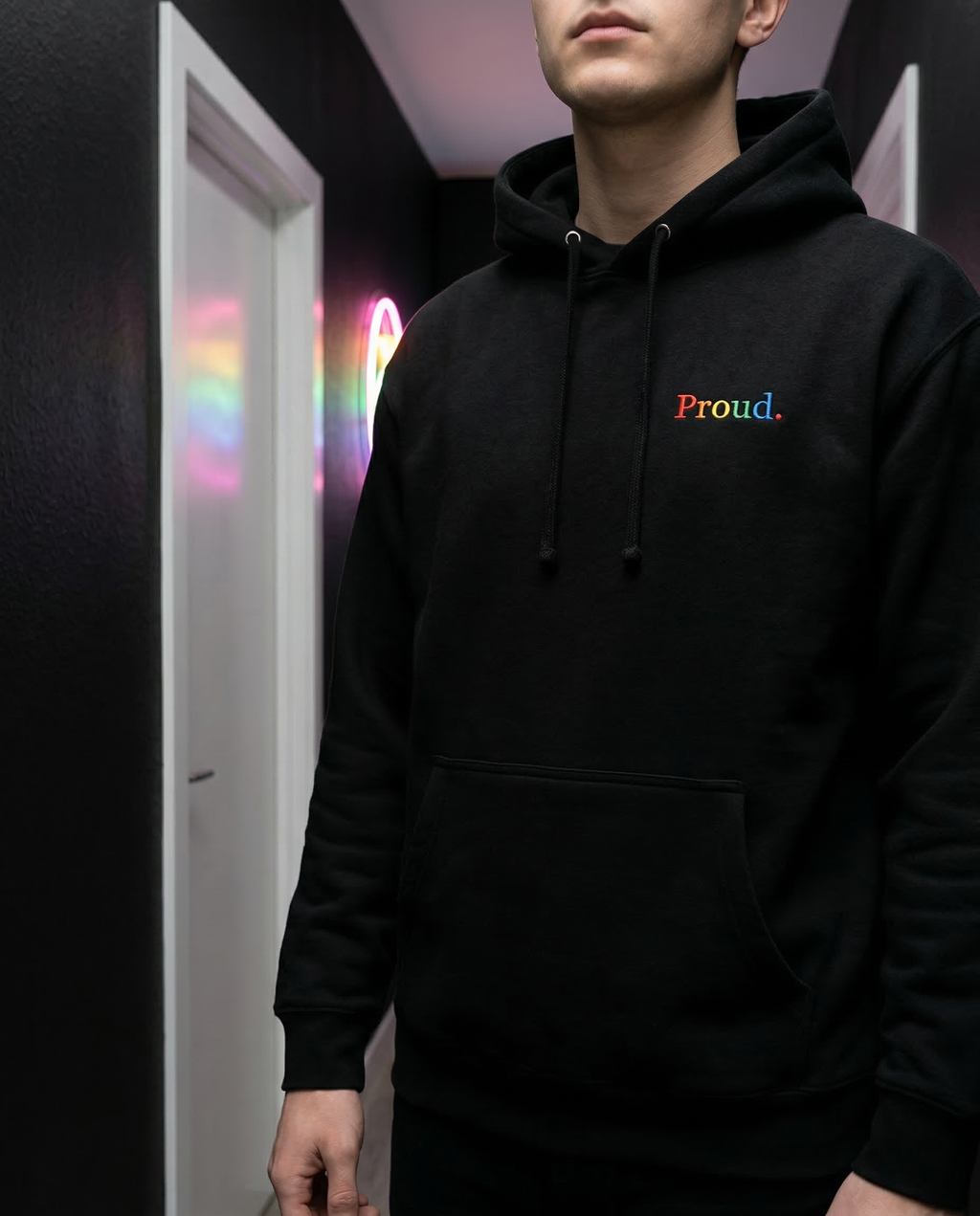 Person wearing a black hoodie with 'Proud.' text in rainbow colors, standing in a room with colorful lights. Proud Premium Pullover Hoodie